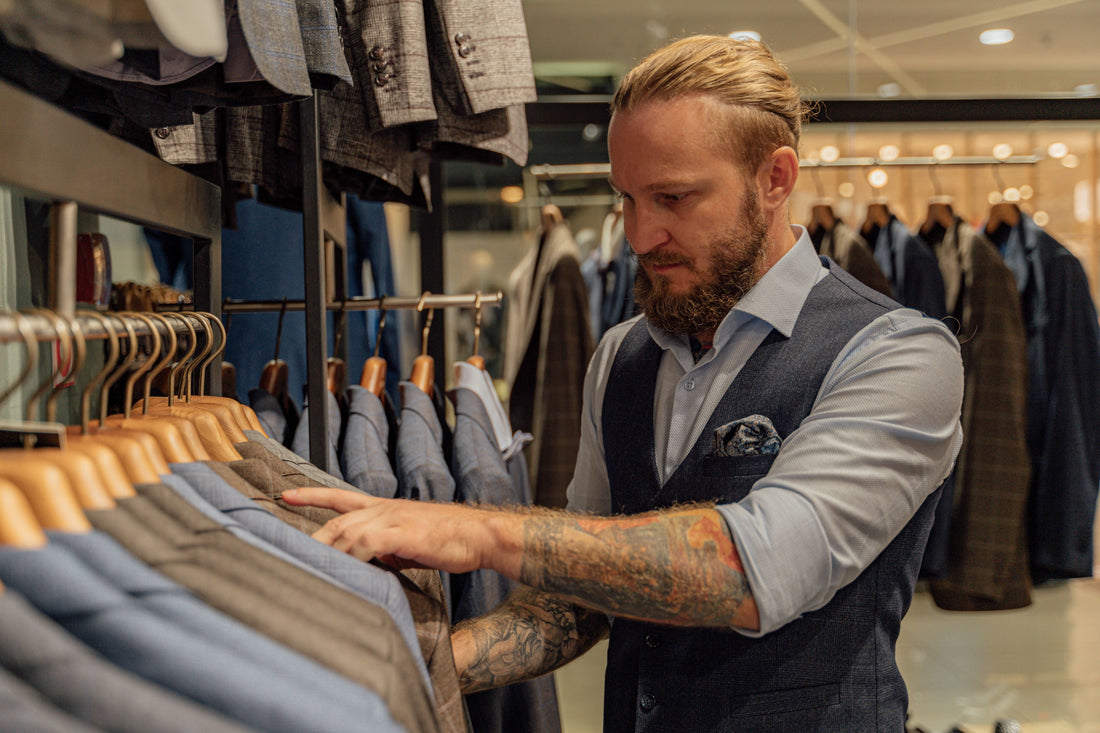 Bearded man with tattooed arms and a man bun browses a suit rack in a high-end menswear store, wearing a tailored vest and blue button-down shirt.