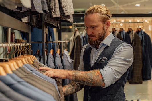 Bearded man with tattooed arms and a man bun browses a suit rack in a high-end menswear store, wearing a tailored vest and blue button-down shirt.